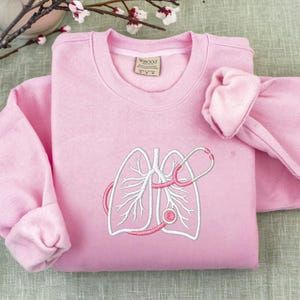 May include: A light pink sweatshirt with a white embroidered lung design and a pink stethoscope. The sweatshirt has long sleeves and a crew neck. The design is centered on the front of the shirt.