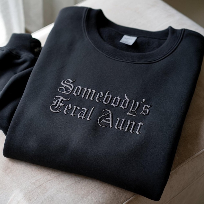 May include: A black sweatshirt with the embroidered text 'Somebody's Feral Aunt' in a gothic-style font. The sweatshirt is folded, with the text prominently displayed. The fabric appears soft and the overall aesthetic is casual and comfortable. The garment is likely a novelty item or a gift.