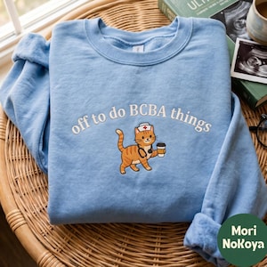 Puede incluir: Una sudadera azul claro de cuello redondo con texto bordado en blanco que dice off to do BCBA things. Debajo del texto hay un gato atigrado naranja bordado con un gorro de enfermero blanco con una cruz roja, un estetoscopio y una taza de café.
