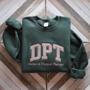 May include: A dark green sweatshirt with the embroidered letters "DPT" in a light tan colour. Below the letters, the words "Doctor of Physical Therapy" are embroidered in a smaller font. The sweatshirt is folded on a wooden surface.