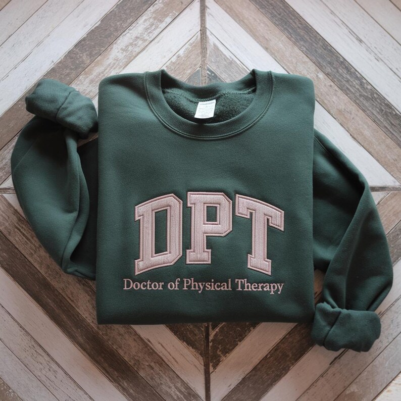 May include: A dark green sweatshirt with the embroidered letters "DPT" in a light tan colour. Below the letters, the words "Doctor of Physical Therapy" are embroidered in a smaller font. The sweatshirt is folded on a wooden surface.