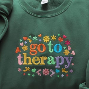May include: A dark green sweatshirt with the embroidered phrase "go to therapy" in rainbow colors. The text is surrounded by colorful embroidered flowers, hearts, and stars.