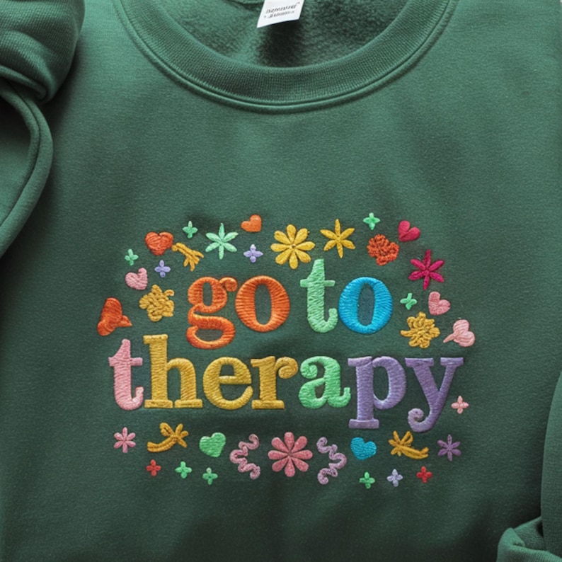 May include: A dark green sweatshirt with the embroidered phrase "go to therapy" in rainbow colors. The text is surrounded by colorful embroidered flowers, hearts, and stars.