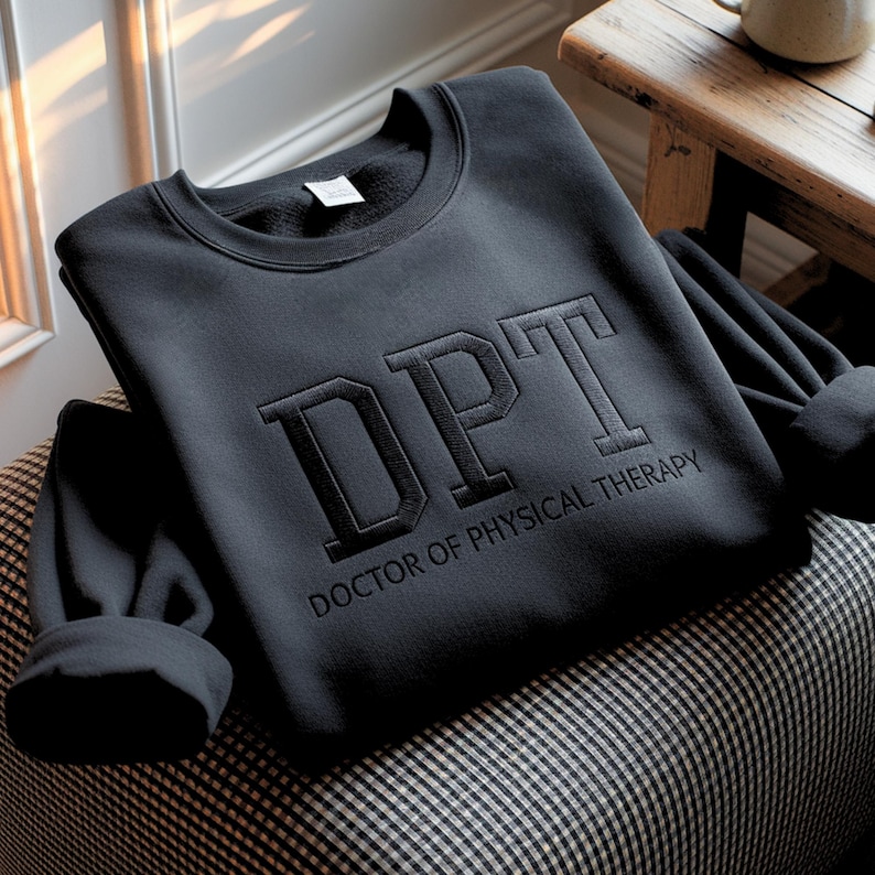 May include: Black sweatshirt with the letters "DPT" embroidered on the front, with "DOCTOR OF PHYSICAL THERAPY" below. The sweatshirt is folded on a black and white checkered patterned chair.