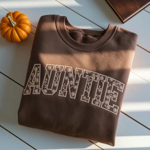 May include: A brown sweatshirt with the embroidered word "AUNTIE" in a floral design. A small orange pumpkin is in the upper left corner. The sweatshirt is folded on a white wooden surface.