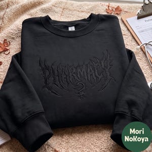 May include: A black crewneck sweatshirt featuring the word PHARMACY embroidered in a dark, gothic-style font across the chest. Below the text is a small, matching black embroidered symbol of a snake coiled around a chalice, known as the Rod of Asclepius.