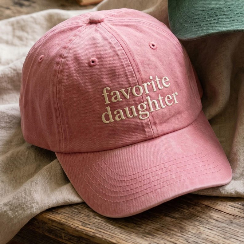 以下が含まれることがあります： 白の「favorite daughter」の刺繍が入った、くすんだピンク色の野球帽。つばがカーブしており、上部に布で覆われたボタンが付いています。柔らかく、質感のある素材で作られています。