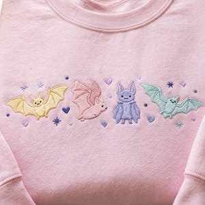 May include: A light pink sweatshirt featuring an embroidered design of four bats in pastel yellow, pink, purple, and light blue. The embroidery includes small purple hearts, dots, and starburst shapes scattered around the bats on the chest area.