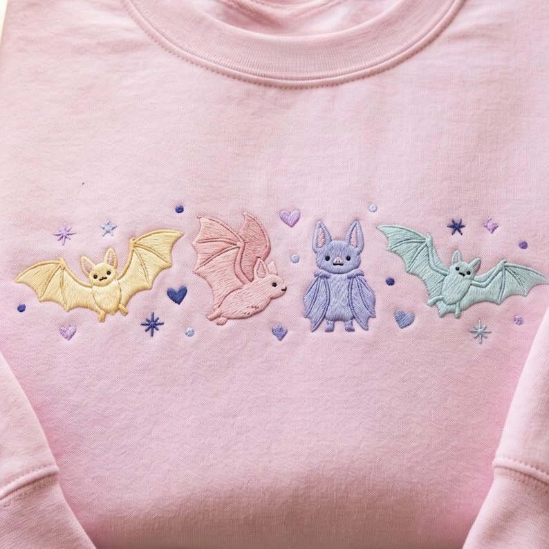 May include: A light pink sweatshirt featuring an embroidered design of four bats in pastel yellow, pink, purple, and light blue. The embroidery includes small purple hearts, dots, and starburst shapes scattered around the bats on the chest area.