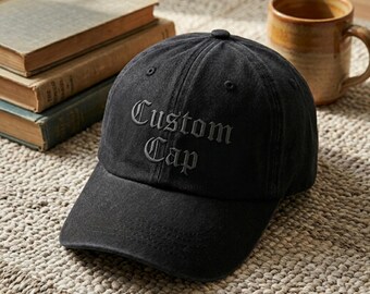 Puede incluir: Una gorra de béisbol negra con un acabado desgastado y lavado. La parte frontal de la gorra presenta el texto "Custom Cap" bordado en una fuente oscura de estilo gótico. La gorra tiene una corona estructurada, una visera curva y ojales.