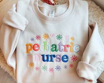 Puede incluir: Sudadera de color crema con las palabras "pediatric nurse" bordadas en letras coloridas. El diseño incluye estrellas y corazones bordados alrededor del texto. La sudadera tiene cuello redondo y mangas largas.