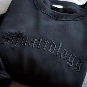 May include: Black crewneck sweatshirt with embroidered text "Stfuattdlaggg" in a gothic font. The sweatshirt is a solid black color and appears to be made of a soft material. The embroidery is in a matching black thread.
