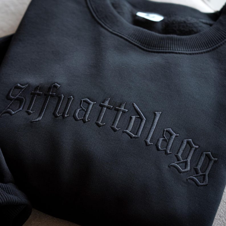 May include: Black crewneck sweatshirt with embroidered text "Stfuattdlaggg" in a gothic font. The sweatshirt is a solid black color and appears to be made of a soft material. The embroidery is in a matching black thread.