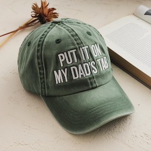 May include: Green baseball cap with the embroidered text "PUT IT ON MY DAD'S TAB" in white. The cap has a curved brim and a distressed look. A book and dried flower are in the background.