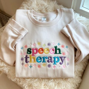 Puede incluir: Una sudadera de cuello redondo color crema con el texto bordado "speech therapy" en letras de bloque multicolores. El diseño está rodeado por pequeñas formas bordadas de estrellas y flores en tonos rosa, azul, amarillo y morado.