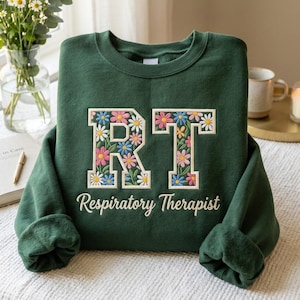 May include: A dark green sweatshirt with the letters "RT" in a floral design. The letters are filled with colorful daisies. Below the letters, the words "Respiratory Therapist" are embroidered in a white script.