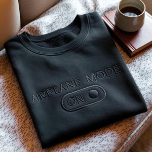 Puede incluir: Sudadera negra de cuello redondo con la inscripci&oacute;n bordada "AIRPLANE MODE ON" y un dise&ntilde;o de interruptor de encendido/apagado. El bordado oscuro coincide con el color de la sudadera. La prenda se muestra sobre una superficie estampada.