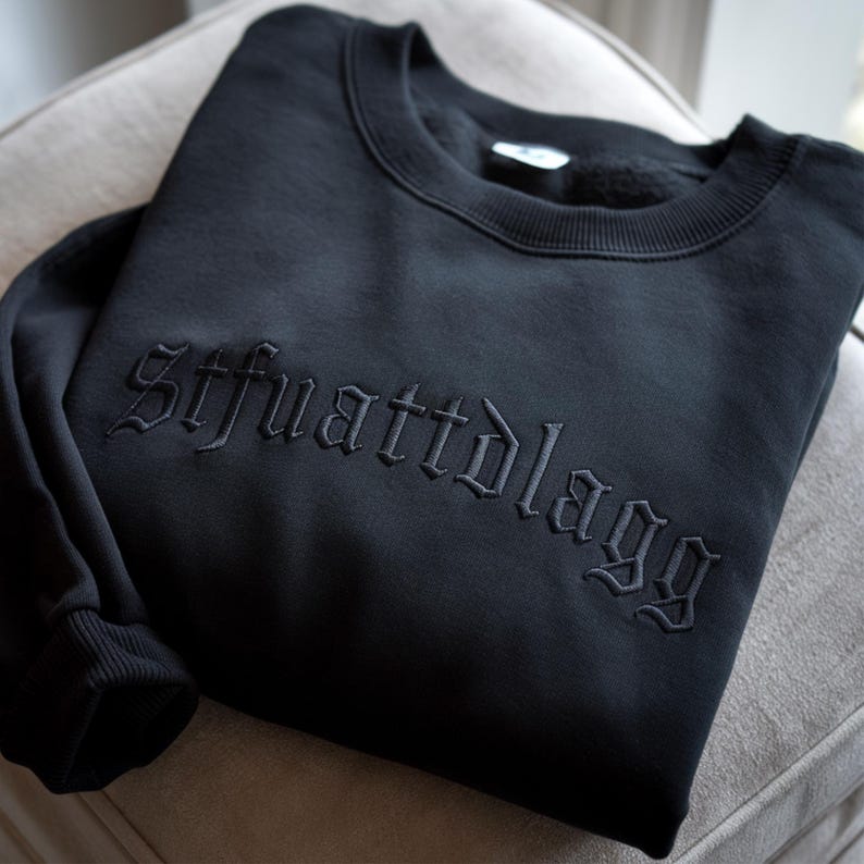 May include: Black sweatshirt with the embroidered text "Sifuattdlagg" in a gothic font. The sweatshirt is folded and resting on a neutral-toned surface. The cuffs and neckline are ribbed.