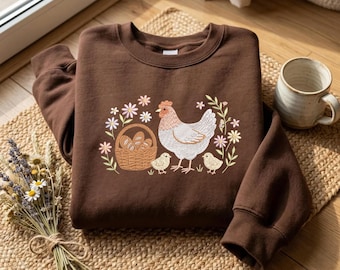May include: A dark brown crewneck sweatshirt featuring an embroidered graphic of a white hen, two small chicks, a woven basket filled with eggs, and various colourful flowers arranged in a circular pattern on the centre of the chest.