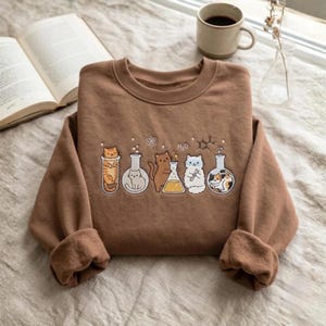 Puede incluir: Una sudadera marrón de cuello redondo con un estampado gráfico de cinco matraces de laboratorio de cristal que contienen varios gatos y un líquido amarillo. El diseño incluye pequeños símbolos científicos como H2O y un icono de átomo.