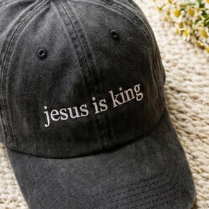 May include: A charcoal gray baseball cap with the embroidered text "jesus is king" in white. The cap has a curved brim and a classic six-panel design. The hat is made of a soft, worn fabric.