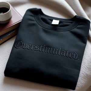 Puede incluir: Sudadera negra de cuello redondo con la palabra "Overstimulated" bordada en fuente g&oacute;tica. La sudadera est&aacute; doblada sobre una superficie de color neutro, con una taza de caf&eacute; y libros en el fondo.