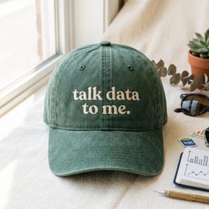 May include: A green baseball cap with the phrase "talk data to me" embroidered in white. The cap is made of a soft, worn material and has a curved brim. The hat is sitting on a white surface.
