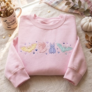 May include: A light pink crewneck sweatshirt featuring an embroidered row of four bats in pastel yellow, pink, purple, and light blue, accented with small embroidered stars and hearts. The fabric is a solid light pink cotton blend material.