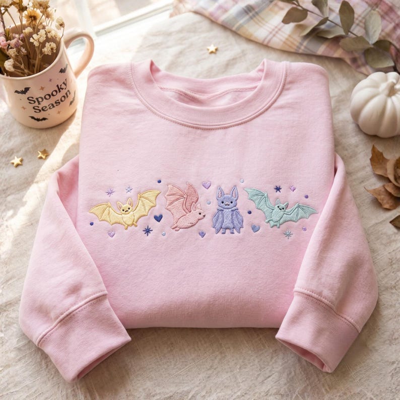 May include: A light pink crewneck sweatshirt featuring an embroidered row of four bats in pastel yellow, pink, purple, and light blue, accented with small embroidered stars and hearts. The fabric is a solid light pink cotton blend material.