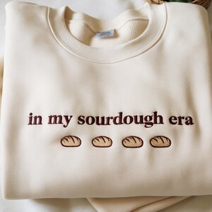 Może przedstawiać: Kremowa bluza z haftowanym napisem "in my sourdough era" brązową nicią. Poniżej tekstu znajdują się cztery haftowane wzory bochenk&oacute;w chleba. Bluza ma okrągły dekolt i luźny kr&oacute;j.
