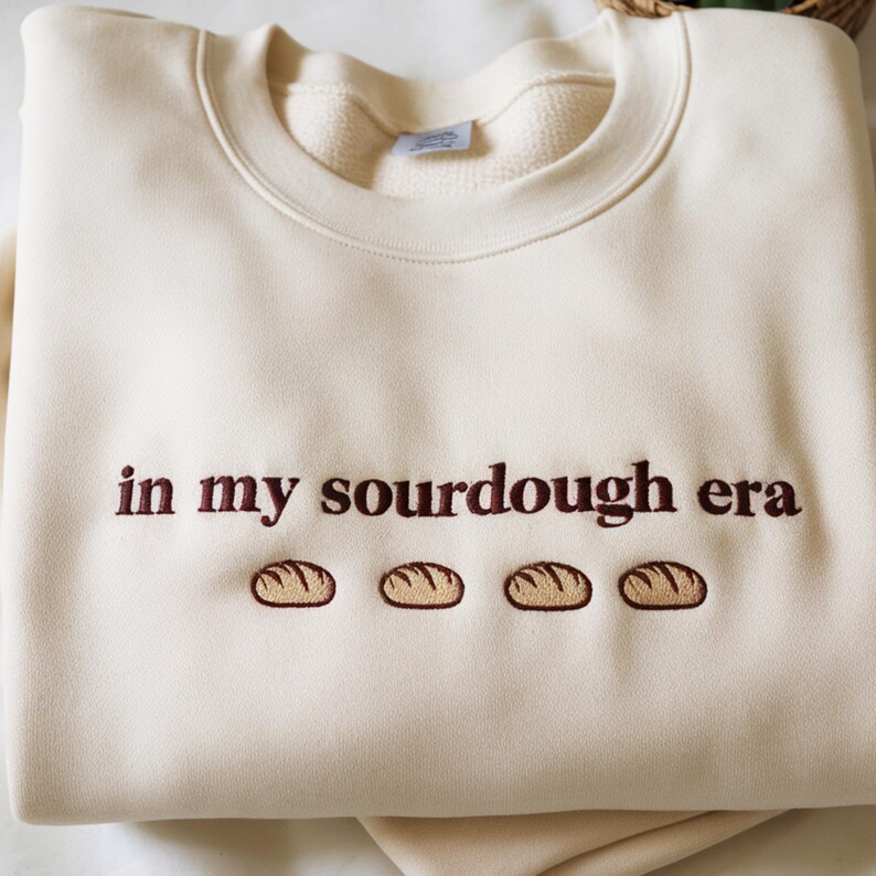 Może przedstawiać: Kremowa bluza z haftowanym napisem "in my sourdough era" brązową nicią. Poniżej tekstu znajdują się cztery haftowane wzory bochenk&oacute;w chleba. Bluza ma okrągły dekolt i luźny kr&oacute;j.