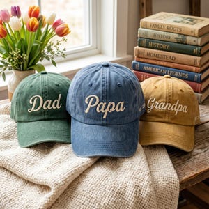 May include: Three baseball caps in green, blue, and tan. Each cap has white embroidered text: "Dad", "Papa", and "Grandpa". The caps are on a cream-colored blanket, with a vase of flowers and a stack of books in the background.