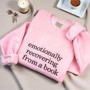 May include: A light pink crewneck sweatshirt featuring the black printed text "emotionally recovering from a book" on the front. The garment has long sleeves with ribbed cuffs and a ribbed neckline, showcasing a simple and clean design.