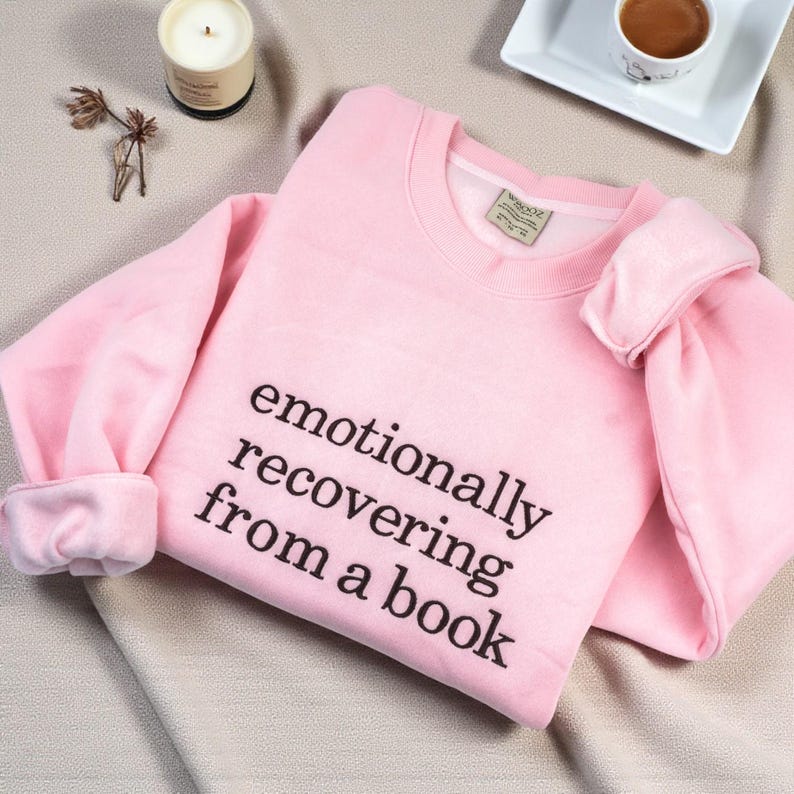May include: A light pink crewneck sweatshirt featuring the black printed text "emotionally recovering from a book" on the front. The garment has long sleeves with ribbed cuffs and a ribbed neckline, showcasing a simple and clean design.