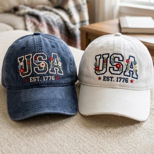 May include: Two baseball caps, one navy blue and one white. Both caps feature the word "USA" in a floral embroidered design, with "EST. 1776" below. The navy cap has a slightly faded look, while the white cap is crisp and clean. Both have a curved brim.