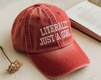 Puede incluir: Gorra de béisbol roja con el texto blanco "LITERALLY JUST A GIRL". La gorra tiene una visera curva y un aspecto ligeramente descolorido. Un libro y una flor seca están en el fondo.