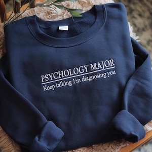 May include: A navy blue crewneck sweatshirt featuring white embroidered text that reads PSYCHOLOGY MAJOR with a horizontal line underneath and the phrase Keep talking I'm diagnosing you below it. The garment has long sleeves and a ribbed collar.