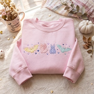 May include: A light pink crewneck sweatshirt featuring an embroidered design across the chest. The embroidery depicts four small bats in pastel yellow, pink, purple, and mint green, accented with tiny purple hearts and star-shaped sparkles.