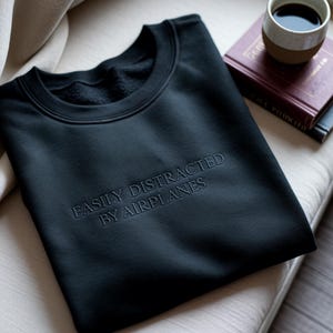 May include: Black crew neck sweatshirt with the embroidered text "EASILY DISTRACTED BY AIRPLANES". The sweatshirt is folded on a light-colored cushion. A cup of coffee and a book are in the background.