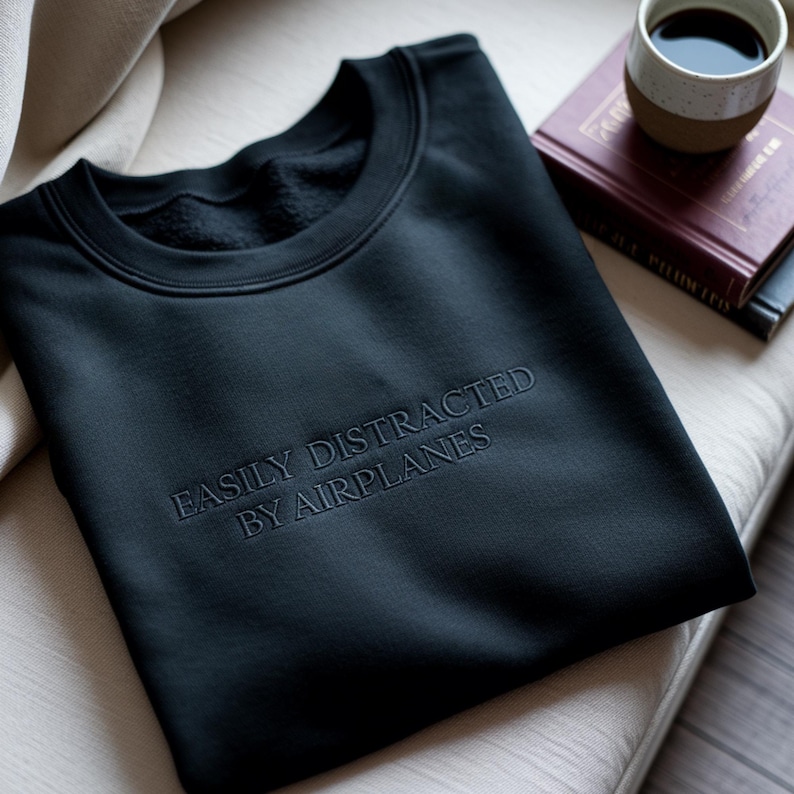 May include: Black crew neck sweatshirt with the embroidered text "EASILY DISTRACTED BY AIRPLANES". The sweatshirt is folded on a light-colored cushion. A cup of coffee and a book are in the background.