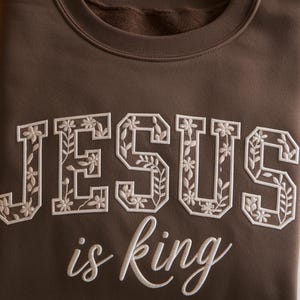 May include: A brown sweatshirt with the words "JESUS is king" embroidered in white. The letters are outlined with a floral design. The sweatshirt has a crew neck and a soft, comfortable appearance.