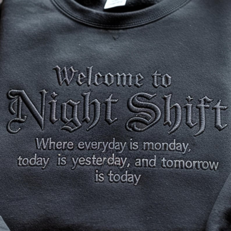 May include: A black crewneck sweatshirt featuring dark gray embroidery. The text reads: Welcome to Night Shift, Where everyday is monday, today is yesterday, and tomorrow is today. The lettering uses a gothic-style font for the main title.