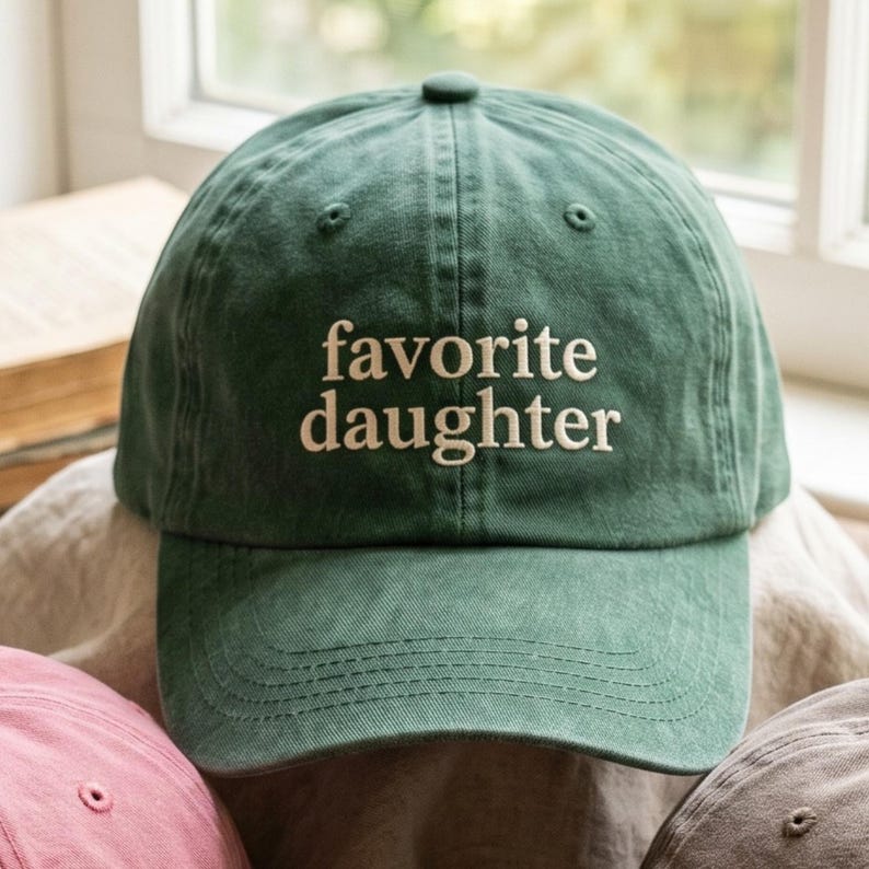 以下が含まれることがあります： 白い文字で「favorite daughter」と刺繍された緑色の野球帽。 つばがカーブしており、上部にボタンが付いています。 ピンクとグレーの他の帽子も部分的に見えています。