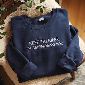 May include: A navy blue sweatshirt with the white text "KEEP TALKING. I'M DIAGNOSING YOU." The garment is folded on a cream-coloured, textured surface. Green leaves are visible in the background.