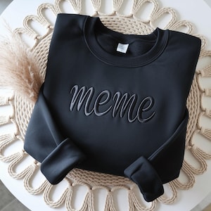 May include: A black long-sleeve sweatshirt featuring the word meme embroidered in a dark grey cursive font across the chest. The garment has a crew neckline, ribbed cuffs, and a white interior neck label visible at the collar area.