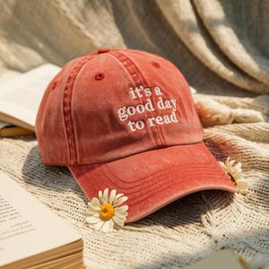 May include: A red baseball cap with the embroidered text "it's a good day to read." The cap has a curved brim and a distressed look. A white daisy with a yellow center is placed on the brim.