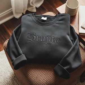 May include: A dark gray sweatshirt with the word "Brujita" embroidered in a gothic font across the chest. The sweatshirt has a crew neck and long sleeves with folded cuffs. The garment is displayed on a woven chair.