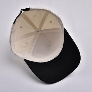 Op de afbeelding: Een baseballpet met een zwarte klep en een cr&egrave;mekleurige kroon. De binnenkant van de pet is cr&egrave;mekleurig met een zwarte verstelbare band. De pet wordt van binnenuit bekeken.