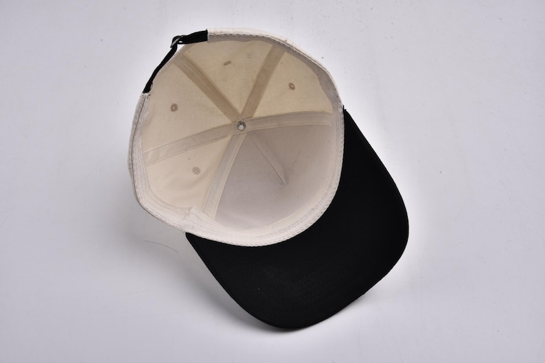 Op de afbeelding: Een baseballpet met een zwarte klep en een cr&egrave;mekleurige kroon. De binnenkant van de pet is cr&egrave;mekleurig met een zwarte verstelbare band. De pet wordt van binnenuit bekeken.