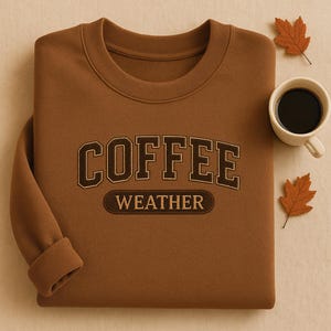 May include: A brown sweatshirt with the embroidered text "COFFEE WEATHER" in a collegiate style. A white coffee cup filled with black coffee and two autumn leaves are placed next to the sweatshirt. The sweatshirt has a crew neck and cuffed sleeves.